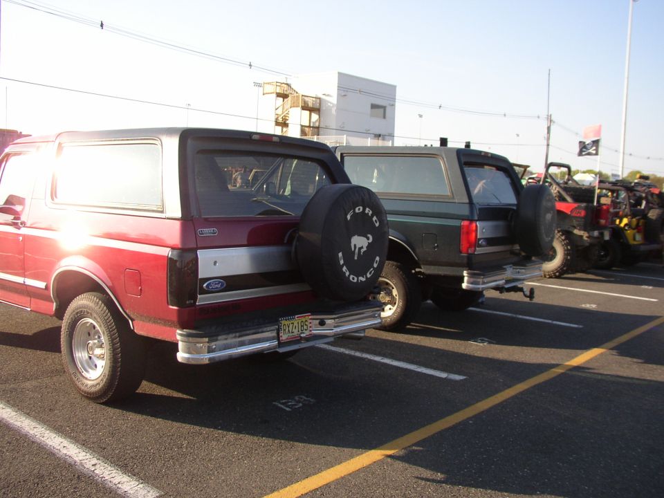 Two Ford Broncos parked side-by-side, one red and one dark green. Both have spare tires mounted on the rear