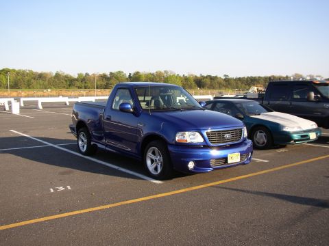 1995 Ford Bronco - A blue Ford F-150 Lightning pickup truck is parked in a lot with other vehicles under a clear sky.
