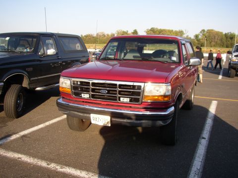 1995 Ford Bronco - Front three-quarter view of a red Ford Bronco parked in a paved lot, showing its chrome-surround egg-crate grille with blue Ford oval, chro…