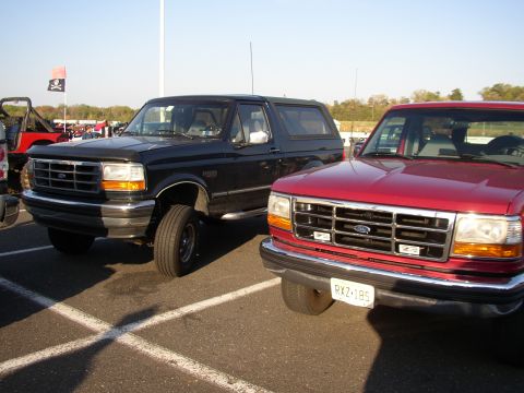 1995 Ford Bronco - Two full-size Ford Broncos parked nose-out in a paved lot at a track or show: a black one on steel wheels with black grille and BRONCO fend…