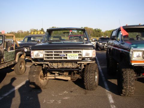 1995 Ford Bronco - Several lifted trucks are parked in a lot under a clear sky, with the front of a black Ford Bronco prominently featured.