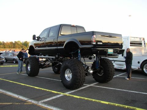 1995 Ford Bronco - A heavily modified black pickup truck with massive tires dominates the scene at a car event.