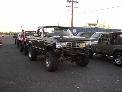 1995 Ford Bronco - A line of lifted trucks and Jeeps are parked at an outdoor event.
