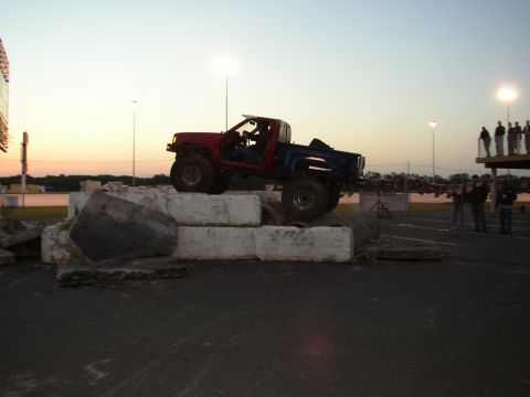 1995 Ford Bronco - A modified pickup truck navigates an obstacle course at dusk, with spectators watching from a platform.