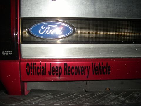 1995 Ford Bronco - Night close-up of a red Ford tailgate showing the blue oval emblem on a brushed-aluminum trim panel, with a black vinyl decal below reading…
