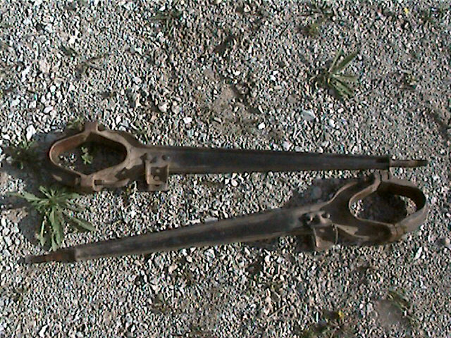 Two rusty metal suspension parts lying on gravel. One part is above the other