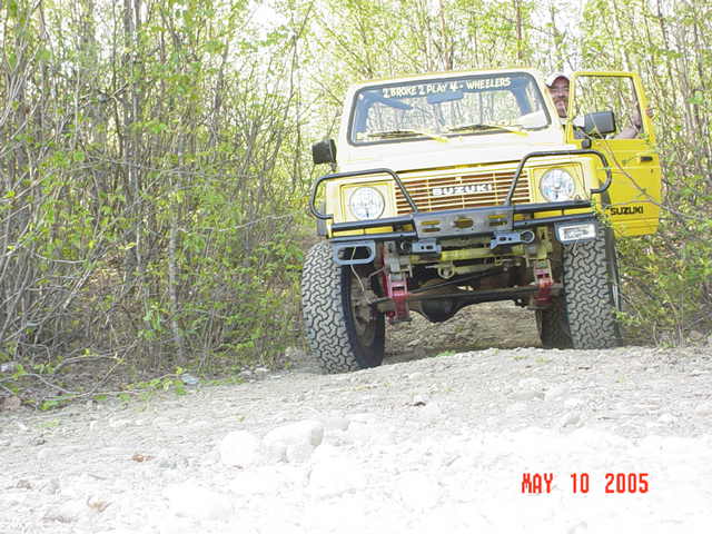 Yellow Suzuki Samurai with brush guard and lift on rocky off-road trail, driver's door open, windshield banner text