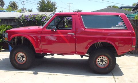 1990 Ford Bronco - red lifted full-size Ford Bronco with a removable hardtop and beadlock-style wheels sits in profile in a residential driveway.