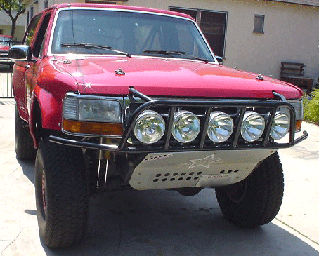 1990 Ford Bronco - Front three-quarter view of a red lifted Ford Bronco with a tube prerunner bumper, five driving lights, and a skid plate.