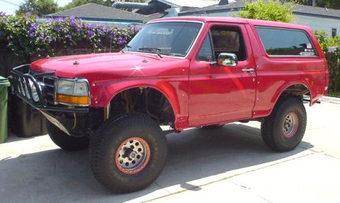 1990 Ford Bronco - red lifted Ford Bronco with a hardtop, tubular prerunner bumper, and large tires parked in a driveway by a flowering hedge.
