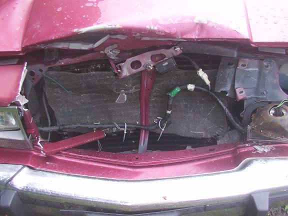 Crushed front end of a maroon sedan with grille removed, showing damaged hood, radiator area, and wiring