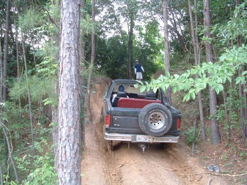 1993 Ford Bronco - A Ford Bronco navigates a muddy trail through a wooded area, with a person standing on the rear tailgate.