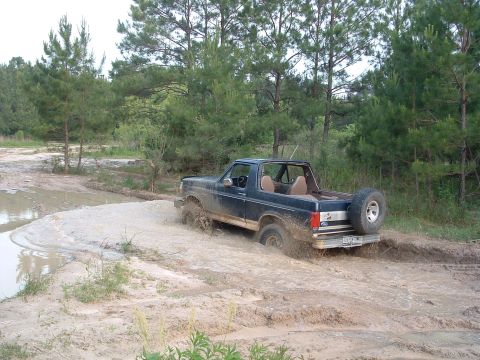 1993 Ford Bronco - A dark blue Ford Bronco navigates a muddy trail, kicking up water and dirt.