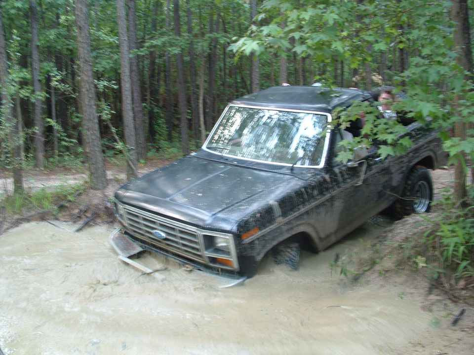 Black Ford Bronco stuck in a muddy ditch in a wooded area. The front end is submerged in water