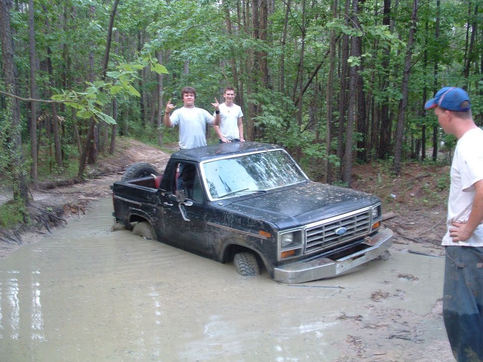 Black Ford Bronco stuck in a mud pit with two people on top and one person watching