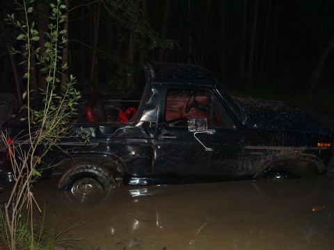 1993 Ford Bronco - A black Ford Bronco is partially submerged in muddy water in a wooded area at night.