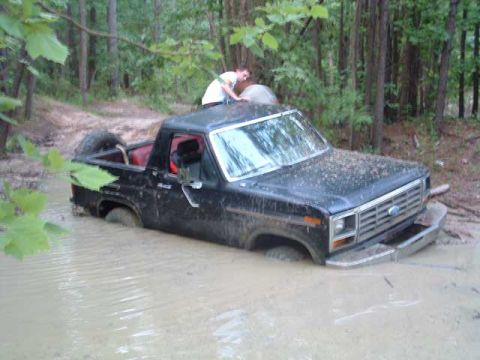 1993 Ford Bronco - A person stands on the roof of a black Ford pickup truck submerged in muddy water in a wooded area.