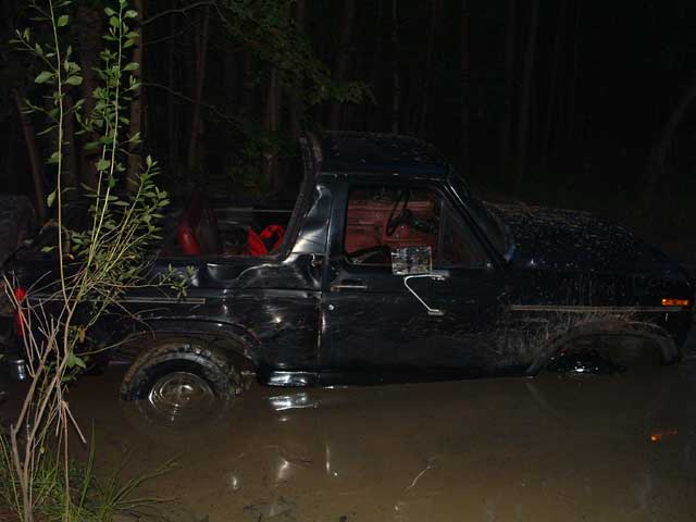 Black topless Ford Bronco sunk to the rockers in muddy water at night in the woods