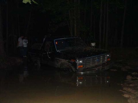 1993 Ford Bronco - A dark Ford pickup truck is stuck in a muddy puddle at night, with trees in the background.