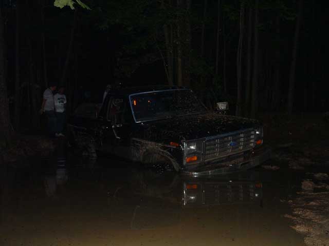 Dark Ford truck stuck up to its rockers in a muddy water hole at night in the woods