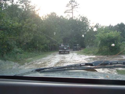 1993 Ford Bronco - Two SUVs navigate a wet, muddy path surrounded by trees, suggesting off-road conditions.