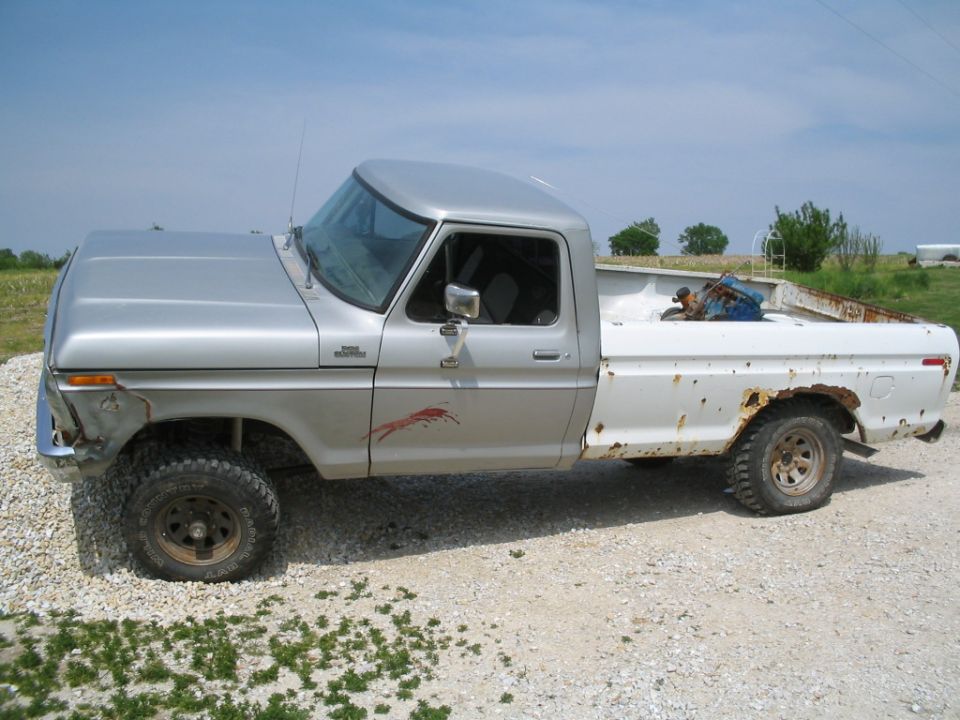 Silver and white 70s Ford pickup with lift and mud tires on gravel in a field