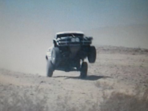 1990 Ford Bronco - off-road race truck catches air over a dirt berm in an open desert, trailing a cloud of dust.