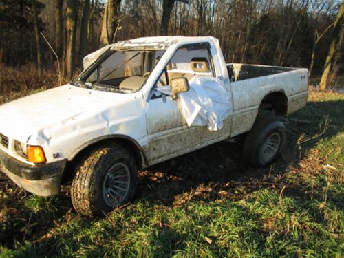 Damaged white compact pickup with crushed roof and missing windshield parked in a grassy field