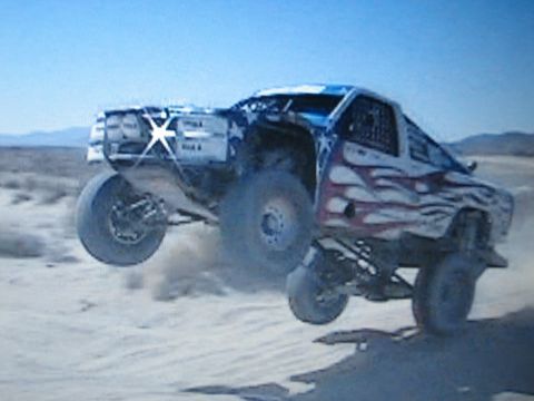 1990 Ford Bronco - blue-and-white off-road race truck with flame graphics and roof light bar leaps over a sandy desert rise.