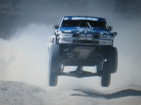 1990 Ford Bronco - blue off-road race truck flies front-on through a cloud of dust with all wheels off the ground.