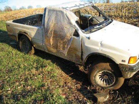 1979 Ford F-150 - This damaged white pickup truck appears stuck in a muddy field, with a tarp obscuring the driver's side door and window.