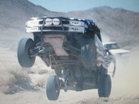 1990 Ford Bronco - off-road race truck jumps high in the desert, exposing its undercarriage and a roof light bar with PIAA-branded lamps.