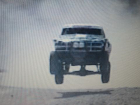 1990 Ford Bronco - light-colored off-road race truck catches air front-on over a desert track in a haze of dust.