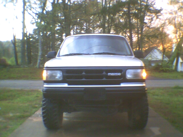 White SUV, front view, headlights on, parked on driveway among trees