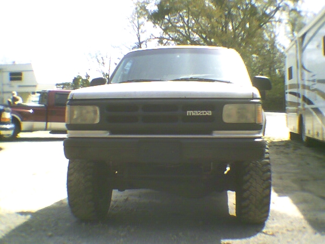 Front view of white/dark Mazda SUV lifted on large tires, parked on gravel