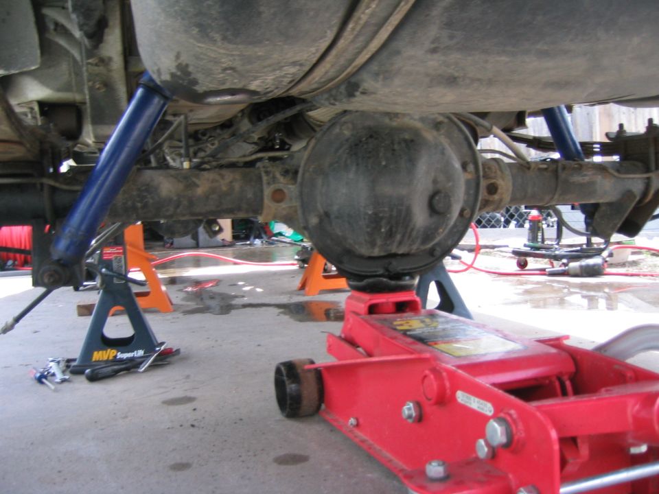 Underside of a vehicle being worked on, showing the rear axle, differential, and suspension components supported by jack stands and a floor jack