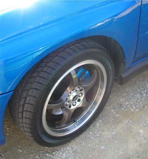 2002 Subaru Impreza - A close-up view of the front wheel and fender of a blue 2002 Subaru Impreza WRX, showing aftermarket wheels and blue brake calipers.