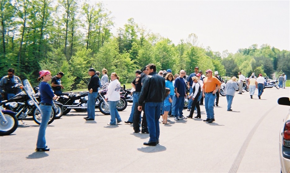 Group of people gathered around motorcycles in a parking lot with trees in the background