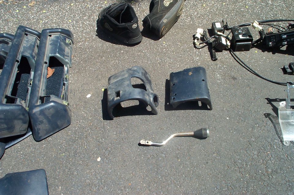 Various car parts including dashboard trim pieces, a steering column cover, and a shift lever laid out on asphalt