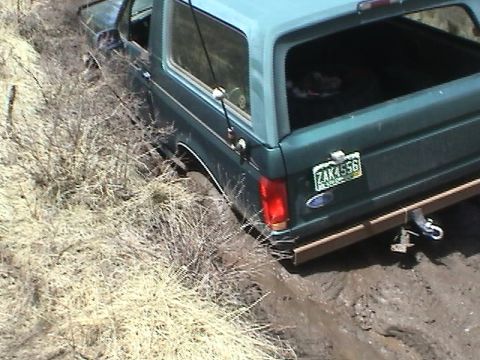 1979 Ford Bronco - Teal-green Ford full-size Bronco stuck nose-down in a muddy rut on a dry brush hillside, photographed from above the rear three-quarter. A…