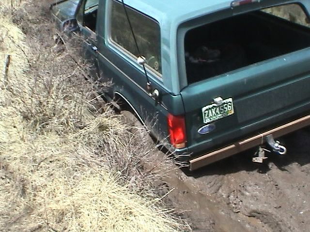 Green Ford Bronco stuck in mud on hillside with board under rear bumper and recovery strap