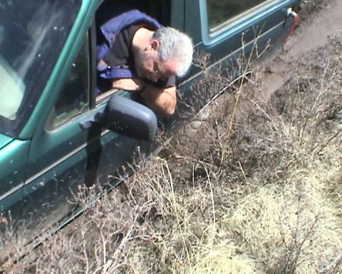 1979 Ford Bronco - A gray-haired man leans out the driver's window of a teal-green truck to look down at the ground as the vehicle sits bogged in a rutted dir…