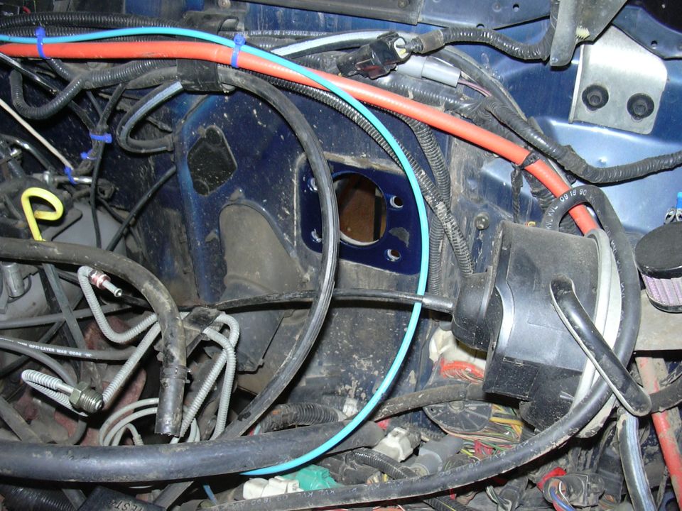 Engine bay with blue paint, red and blue hoses, and black wiring. A circular opening is visible in the blue firewall