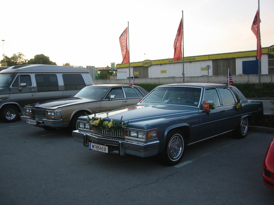 Blue Cadillac sedan with flowers on the hood and American flags on the roof, parked next to a gold sedan and a van
