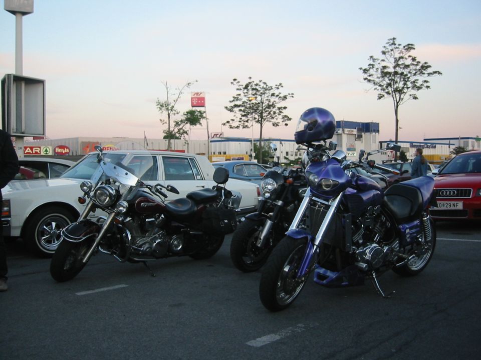 Three motorcycles parked in a lot with cars and trees
