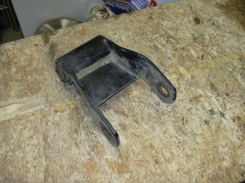 1991 Ford Bronco - A black steel mounting bracket with two bolt holes and a pivot/bushing joint on one end, resting on an oriented strand board (OSB) workbenc…