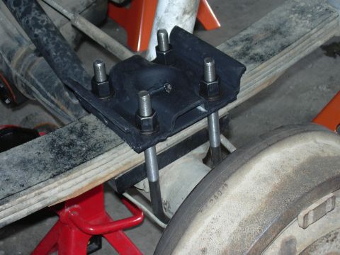 1991 Ford Bronco - Close-up of a black cast metal spring perch or shackle plate mounted over a leaf spring on a vehicle's rear axle, with four vertical thread…