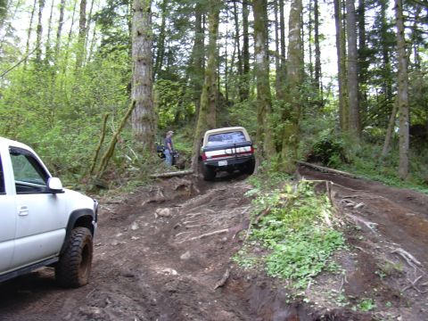 1991 Ford Bronco - Two off-road vehicles tackle a challenging muddy trail through a dense forest.