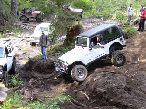 1991 Ford Bronco - Several off-road vehicles traverse a challenging muddy trail in a wooded area.