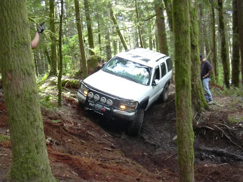 1991 Ford Bronco - A white SUV tackles a challenging muddy incline on an off-road trail surrounded by dense forest.